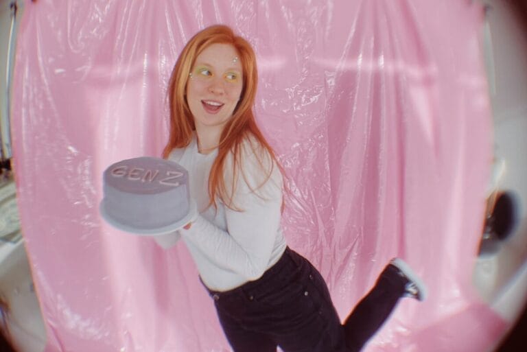 Teenage girl with red hair, white shirt and black pants holding light purple cake that says Gen Z